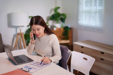 Bir kadın birden fazla iş yapıyor, bilgisayarını kullanırken telefonla konuşuyor ve masadaki belgeleri inceliyor..