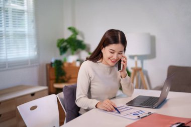 Bir kadın birden fazla iş yapıyor, bilgisayarını kullanırken telefonla konuşuyor ve masadaki belgeleri inceliyor..