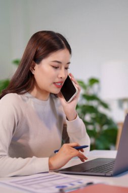 Bir kadın birden fazla iş yapıyor, bilgisayarını kullanırken telefonla konuşuyor ve masadaki belgeleri inceliyor..