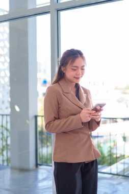 Geniş ve modern bir ofiste akıllı telefonunu kontrol ederken gülümseyen genç Asyalı iş kadını.