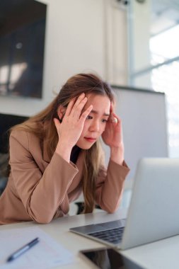 Genç Asyalı iş kadını dizüstü bilgisayarına bakıp iş yerindeki sorunları analiz ederken stresli hissediyor