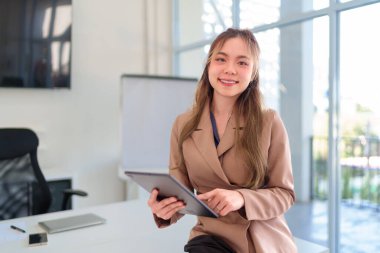 Parlak ve modern ofis ortamında tablet kullanırken gülümseyen genç Asyalı iş kadını
