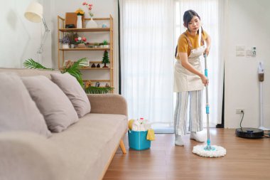 Asyalı hizmetçi, modern oturma odasındaki ahşap zemini temizlik malzemeleriyle fırçalıyor.