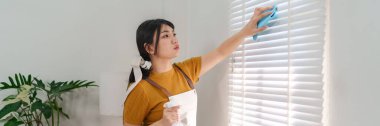 Asyalı hizmetçi modern bir evde sprey şişe ve bezle pencereyi temizliyor.