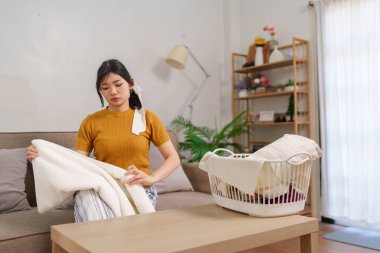 Asyalı hizmetçi, rahat bir oturma odasında çamaşır sepetinin yanında kıyafet katlıyor.