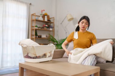 Asyalı hizmetçi, rahat bir oturma odasında çamaşır sepetinin yanında kıyafet katlıyor.
