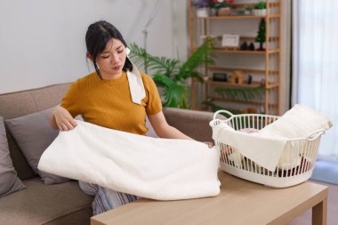 Asyalı hizmetçi, rahat bir oturma odasında çamaşır sepetinin yanında kıyafet katlıyor.