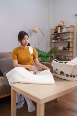 Asyalı hizmetçi, rahat bir oturma odasında çamaşır sepetinin yanında kıyafet katlıyor.