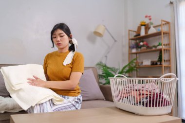 Asyalı hizmetçi, rahat bir oturma odasında çamaşır sepetinin yanında kıyafet katlıyor.