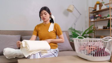 Asyalı hizmetçi, rahat bir oturma odasında çamaşır sepetinin yanında kıyafet katlıyor.