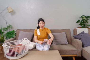 Asyalı hizmetçi, rahat bir oturma odasında çamaşır sepetinin yanında kıyafet katlıyor.