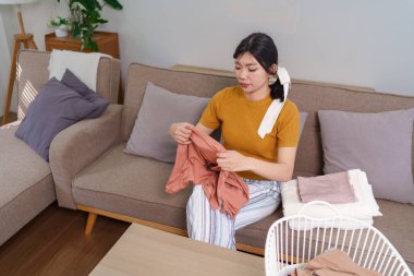 Asyalı hizmetçi, rahat bir oturma odasında çamaşır sepetinin yanında kıyafet katlıyor.