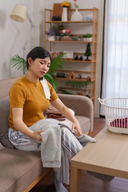 Asyalı hizmetçi, rahat bir oturma odasında çamaşır sepetinin yanında kıyafet katlıyor.