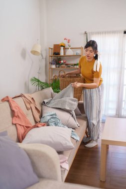Asyalı hizmetçi rahat bir oturma odasında çamaşır sepetine kıyafet topluyor.