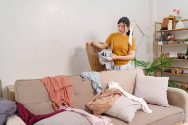 Asyalı hizmetçi rahat bir oturma odasında çamaşır sepetine kıyafet topluyor.