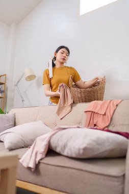 Asyalı hizmetçi rahat bir oturma odasında çamaşır sepetine kıyafet topluyor.