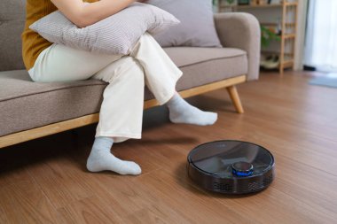 Robot süpürge modern bir oturma odasının zeminini temizlerken Asyalı kadın akıllı telefonunu kullanıyor.