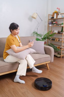 Robot süpürge modern bir oturma odasının zeminini temizlerken Asyalı kadın akıllı telefonunu kullanıyor.