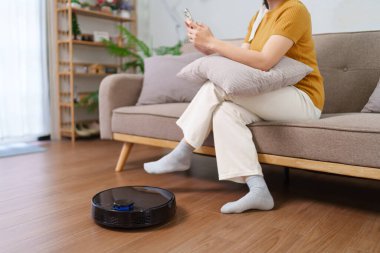 Robot süpürge modern bir oturma odasının zeminini temizlerken Asyalı kadın akıllı telefonunu kullanıyor.