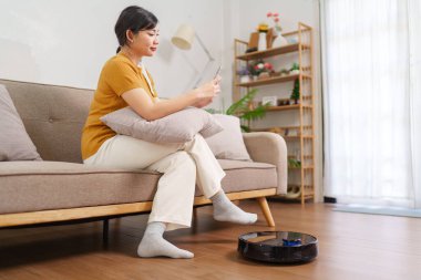 Robot süpürge modern bir oturma odasının zeminini temizlerken Asyalı kadın akıllı telefonunu kullanıyor.