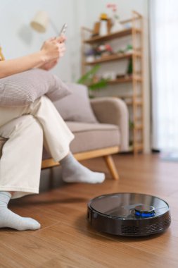 Robot süpürge modern bir oturma odasının zeminini temizlerken Asyalı kadın akıllı telefonunu kullanıyor.