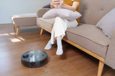 Robot süpürge modern bir oturma odasının zeminini temizlerken Asyalı kadın akıllı telefonunu kullanıyor.
