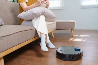 Robot süpürge modern bir oturma odasının zeminini temizlerken Asyalı kadın akıllı telefonunu kullanıyor.