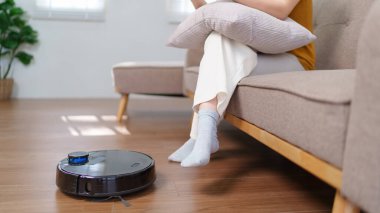 Robot süpürge modern bir oturma odasının zeminini temizlerken Asyalı kadın akıllı telefonunu kullanıyor.