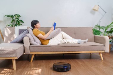 Robot süpürge modern bir oturma odasında yerleri temizlerken Asyalı kadın kitap okuyor.
