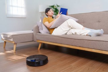 Robot süpürge modern bir oturma odasında yerleri temizlerken Asyalı kadın kitap okuyor.