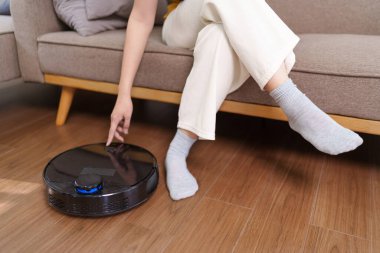 Asyalı kadın rahat bir oturma odasında kanepede otururken robot süpürgesini açıyor.