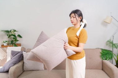 Asyalı hizmetçi rahat bir oturma odasındaki tozu temizlemek için yastıkları sallıyor.