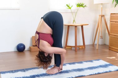 Kıdemli Asyalı kadın Uttanasana çalışıyor. Konforlu bir ev ortamında yoga minderinin üzerinde öne eğiliyor.