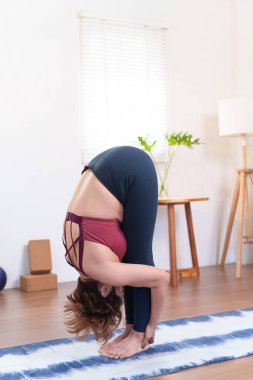 Kıdemli Asyalı kadın Uttanasana çalışıyor. Konforlu bir ev ortamında yoga minderinin üzerinde öne eğiliyor.