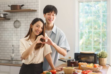Genç çift mutfakta el hareketleriyle taze yiyecek ve doğal ışıkla çevrili.