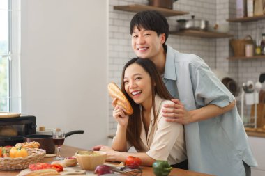 Gülümseyen genç çift, taze yemek ve aşkla dolu modern mutfağa neşeyle poz veriyor.