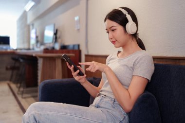 Kanepede oturup kulaklıkla müzik dinleyen ve akıllı telefondan şarkı okuyan genç bir kadın.