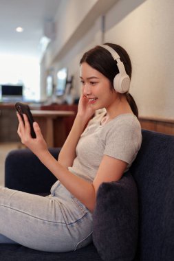 Kanepede oturup kulaklıkla müzik dinleyen ve akıllı telefondan şarkı okuyan genç bir kadın.