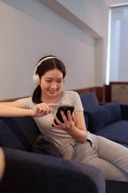 Kanepede oturup kulaklıkla müzik dinleyen ve akıllı telefondan şarkı okuyan genç bir kadın.