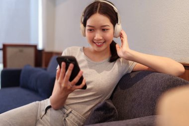 Kanepede oturup kulaklıkla müzik dinleyen ve akıllı telefondan şarkı okuyan genç bir kadın.