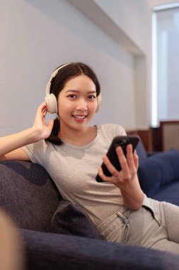 Kanepede oturup kulaklıkla müzik dinleyen ve akıllı telefondan şarkı okuyan genç bir kadın.