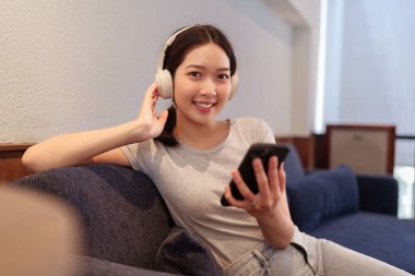 Kanepede oturup kulaklıkla müzik dinleyen ve akıllı telefondan şarkı okuyan genç bir kadın.