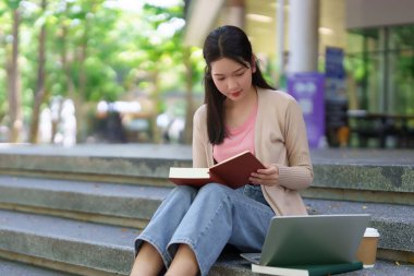 Genç Asyalı kız öğrenci kampüs merdivenlerinde kitap, dizüstü bilgisayar ve kahveyle ders çalışıyor. Eğitim konsepti. Üniversite öğrencisi tatilde kitap okuyor..