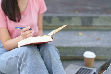 Asyalı genç bir kadın merdivenlerde ders çalışıyor. O bir kalem tutuyor ve yanında bir dizüstü bilgisayar ve bir kahve fincanı olan bir kitap okuyor. Dışarıdaki eğitim hedeflerine odaklanmıştı..