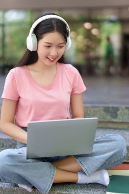 Genç Asyalı kadın kulaklığı açık dizüstü bilgisayar kullanarak oturuyor. Üniversite kampüsünde çevrimiçi çalışıyor, odaklanmış ve gülümsüyor. Daha iyi bir gelecek için eğitim ve e-öğrenim
