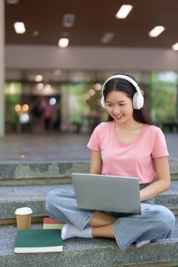 Genç Asyalı kadın kampüste online eğitimden hoşlanıyor. Kulaklık takarak, yanında kahve ve kitap olan bir dizüstü bilgisayar kullanarak verimli bir çalışma seansı yapıyor.