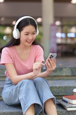 Asyalı genç bir kadın üniversite kampüsünde akıllı telefonunu kullanarak dışarıda kulaklıkla çalışmaktan hoşlanıyor. Eğitimine, yaşam tarzına ve bilgiye odaklan.