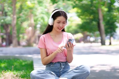 Çevrimiçi eğitim için telefon ve kulaklık kullanan genç bir Asyalı kadın. Parkta müzik ve sosyal medyanın tadını çıkararak yaşam tarzını ve öğrenme deneyimini geliştirdi..