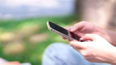 Asyalı bir kadının akıllı telefonu tutarken yakın plan çekimleri. Parkta öğrenme, online iletişim ya da sosyal medya bağlantısı için açık havada kullanılıyor.