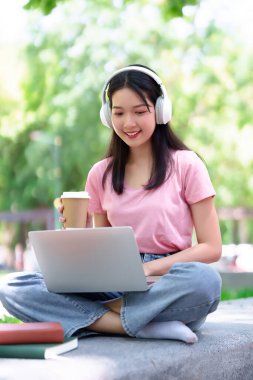 Genç Asyalı kadın dışarıda bilgisayar ve kahveyle çalışıyor. Kız parkta online eğitim için kulaklık kullanıyor. Mutlu öğrenci teknolojiyle e-öğrenmeye odaklanmış..